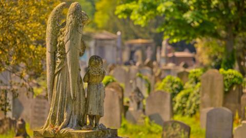 An statue of an angel and a small child stands among Victorian gravestones in a cemetery surrounded by greenery.