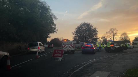 A Y junction with cars queuing in the left lane and multiple police cars and vans parked up on the right hand side behind a red and white road closed sign