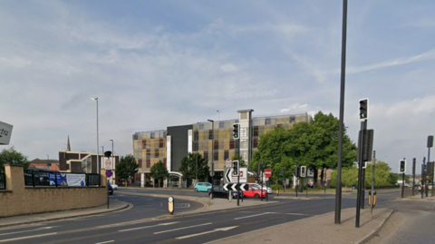 An image of the junction of Centenary Way and Drummond Street. Vehicles are stopped at traffic lights, waiting to turn. A five-storey building is in the background.