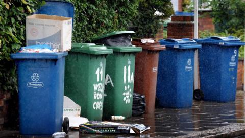 Six waste collection and recycling bins in a row. One is overflowing with rubbish.