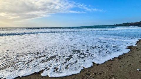 A wave froths up on a sandy beach at Portcatho in Cornwall. The sky above is blue with some cloud visible to the left of frame.