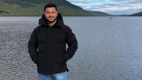 Man in a black jacket and jeans with black hair and beard stands smiling on a jetty in front of a lake with hills and a boat beyond.