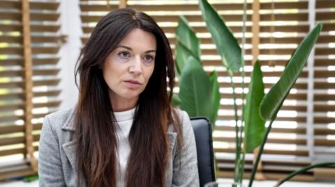 Zoe Trafford has long brown hair and wears a grey blazer and white top underneath. She is sitting against a background of wooden blinds against windows. The leaves of an indoor plant are also there.