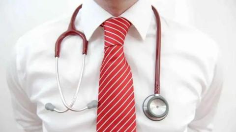 A close-up of a man wearing a white shirt and red and white striped tie, with a stethoscope around his neck.