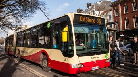 An articulated bus unloading passengers at a bus stop. It is red and yellow with Old Steine and number 25 on the front