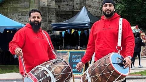 Members of the Northern Dholis who have black hair and beards and are wearing red hoodies who are Bhangra Dhol players. They are each holding a drum. They are smiling and are at a festival with stalls in the background.