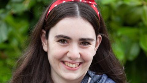 A head-and-shoulders shot of Lucy smiling widely and looking directly at a camera. She has a red and white polka dot bow in her dark hair. She appears to be standing in front of a tree though the background is blurred.