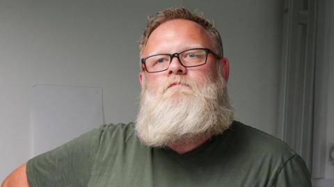 Aaron Clark is a bearded man in glasses. He is stood in a hospital ward.