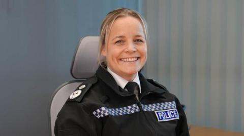 Katy Barrow-Grint is sitting in a desk chair in police uniform. She has long blonde hair and is smiling.