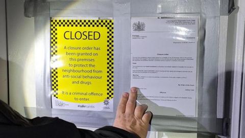 A closure notice placed on a door. The "closed" notice is yellow and there is an explanatory notice taped next to it. A police officer's hand can also be seen, holding the notice on the door