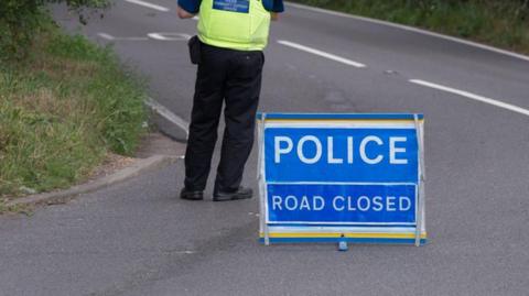 A blue sign of a road closure saying 'police, road closed', with an officer is black trousers stood close by.