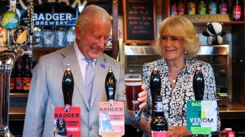 King Charles enjoys a pint next to Queen Camilla in a pub as they both smile