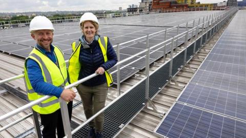 two people in high vis jackets stand on an elevated metal walkway on top of a roof with solar panels 