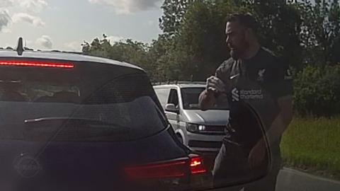 A man with short dark hair and beard looks on at a car angrily on a country road