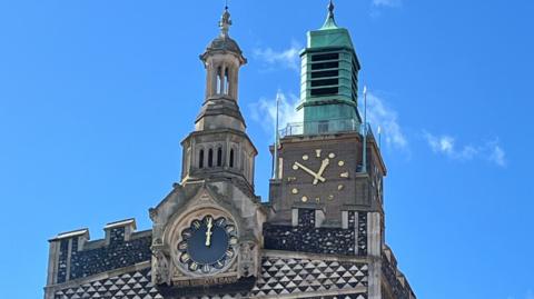 The Norwich Guildhall clock, displaying the time as noon, and City Hall's clock behind it displays the time as 12:50.
