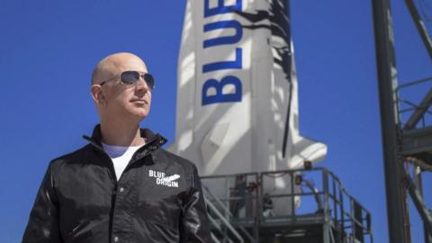 Jeff Bezos wears sunglasses, a white top and a black jacket with the text Blue Origin written on it. Behind him is a white rocket with blue text reading 'Blue'.