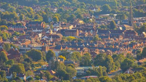 View of Dorking town centre from Surrey Hills. Sun shining on the buildings, in between green trees are lots of houses and an obvious church spire. 