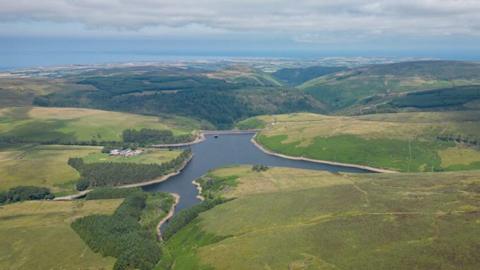 An aerial view on Sulby Reservoir with surrounded by green fields, forest areas and hills. The sea can be seen on the horizon and there is a cloudy blue sky above.