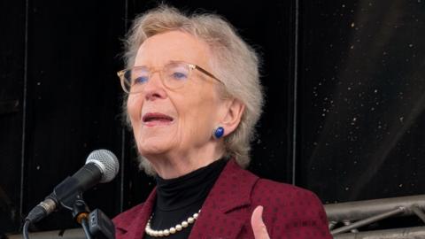 Robinson has short blonde hair and is wearing glasses, blue earrings, a pearl necklace, a black top and red blazer.