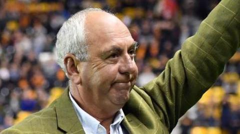 A man with white hair, bald at the front, waves to a football crowd. He is wearing a green jacket with a blue shirt