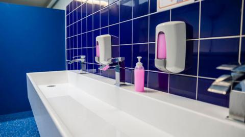 Sinks and soap dispensers in a school toilet with blue tiles 