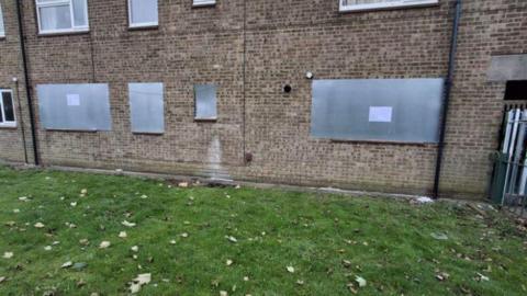 A brick property with its windows closed up by pieces of metal. There is grass with leaves on in front of the property.