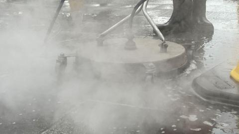 A machine surrounded by steam and water spray is used to get stuck on gum off the pavement. it is surrounded by dirty water.