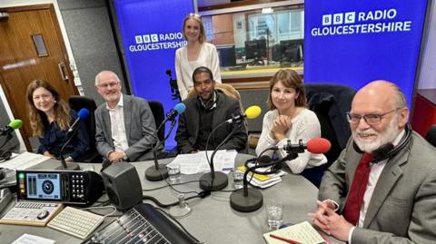 Three men and two women sit in a row along a desk in a radio studio. They all have colourful microphones in front of them. The presenter is standing behind them and BBC Radio Gloucestershire branding can be seen in the background. A few pieces of radio recording equipment, including mixers and speakers, can be seen at the bottom of the image.