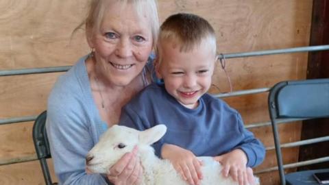 A smiling woman with light blonde hair, wearing a grey cardigan and sat on a black fold-out chair with a young smiling blonde boy wearing a blue jumper on her lap. On his lap is a white baby lamb, and both the woman and boy have their hands on the lamb.