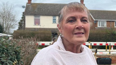 A woman with short hair and wearing a white jumper looking into the camera. Behind her are some hoardings on the road and houses on the other side. She is standing on a driveway which has pebbles on it and a hedge on the left.