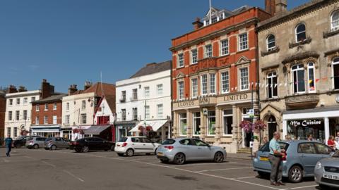 A row of buildings housing shops, including a bank and restaurant, inside historic buildings. There are car parking bays along the road, with about half a dozen cars in them.
