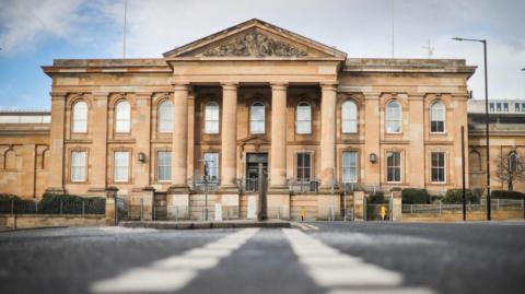 The exterior of Dundee Sheriff Court. The photo is taken from ground level so traffic lines are visible. It is a large sandstone building with four columns at the front.