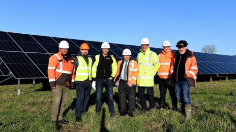 Seven men stood in a field in front of large solar panels. They are all wearing jeans, yellow or orange hi-visibility vests or jackets, and construction hats. The sky is blue.