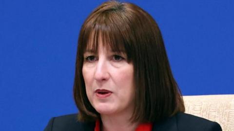 A headshot of Rachel Reeves, with shoulder-length brown hair, wearing a suit jacket and red top, against a blue background at the China-UK Financial Services Summit in Beijing
