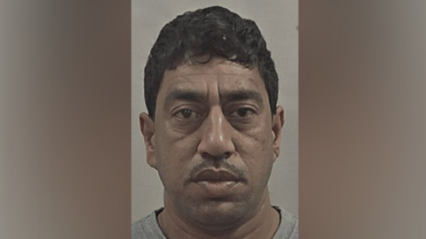 A police custody shot of Malik Shahzad. He has short black hair and a moustache. He is looking at the camera and wearing a grey t-shirt. The wall behind him is grey. 