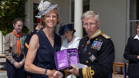 Philippa Stahelin is pictured on the left wearing a dark blue sleeveless dress with a grey hat. She had short hair that ends before her shoulders and is wearing a pearl necklace. The Lieutenant-Governor is positioned on the right and is shaking the hand of Mrs Stahelin while presenting her with a purple velvet lined box containing her award, which is made of glass. He is seen wearing military uniform in a dark blue colour. He has several medals pinned to his uniform. He is also wearing glasses and has short white hair.