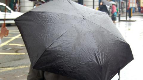 A person stands by a road crossing in a built-up area. They are under a black umbrella.