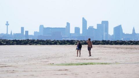 Two people walking on New Brighton Beach on The Wirral, Merseyside. The Liverpool waterfront and skyline is in the distance.