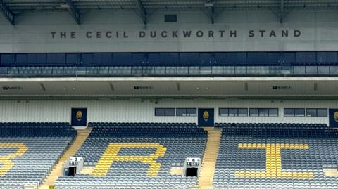 The stand as viewed from the pitch's halfway mark. A large single-tier section of blue seats are shown, with interspersed yellow seats spelling out some of the letters of Warriors - the letters R and I are depicted. Above that, under the stadium roof is the wording The Cecil Duckworth Stand. The wording is in blue, on a white background, and in capital letters.