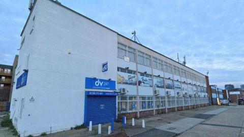 A google image of a large, white, relatively run-down looking commercial building with windows across the facade. There is a blue logo with the wording 'dv 247' on the left hand side of the building.