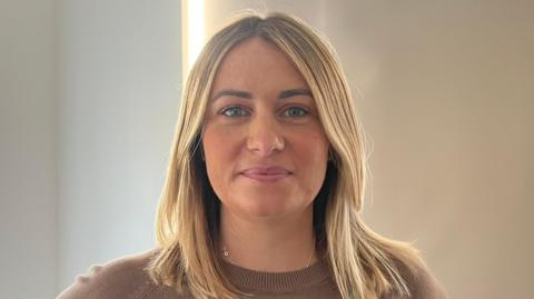 Image of Mrs Madden-Giles in her living room. She has shoulder-length, straight, blonde hair and is wearing a long-sleeved, fitted top in a warm brown shade. The background features a neutral-coloured wall with a tall window covered by a blind. 