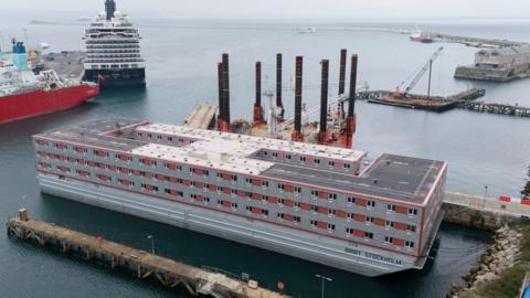 The Bibby Stockholm accommodation barge, with windows on three main floors, is moored alongside a quay at Portland Port. A cruise ship is moored nearby.