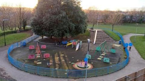Playground in Sheffield with play equipment including a slide and roundabout, and a large tree. A blue fence encircles the playground.