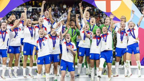 The Lionesses team stand on the podium, each player wearing the England jerseys and a gold medal with their fists in the air in celebration. 