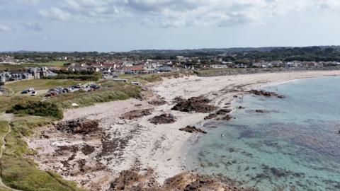 A wide sandy beach next to a light blue sea, with marram grass dunes and houses behind it.