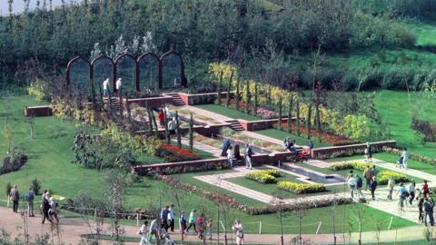 People walk round flower beds on part of the exhibition site. Pathways and colourful flower beds can be seen across a green lawn. 