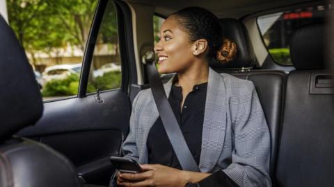 A woman sits in the back of a car with a seatbelt across her body. She has black hair pulled back behind her head and wears a grey jacket with large checks. She has a black top underneath and holds a phone in her hands.