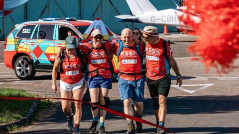 Sophy Dunn, Helene Poursain, Duncan Bootland and Warren Porritt in red KSS shirts walking with backpacks on and their arms around each other. They are walking towards a red ribbon. A KSS vehicle and the tail of a light aircraft can be seen behind them.