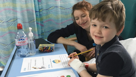 Carter sitting up in a hospital bed doing some school work, with his mum sitting in a chair next to him