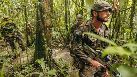 Three Army soldiers dressed in camouflage uniforms and armed with machine guns walk through streams and the greenery of a jungle.